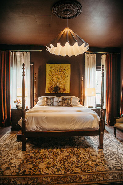 King-size 4-poster bed centered between two windows, dark maroon paint saturates the room with a large modern light fixture over top