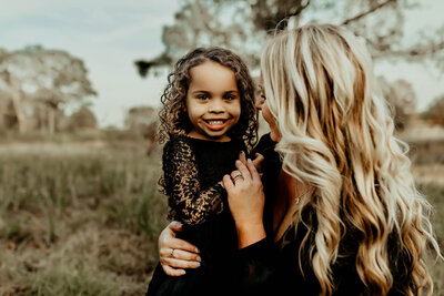 A woman with long, blonde hair holds a smiling child with curly hair in an outdoor setting. Both are wearing black clothing, beautifully captured by Fire Family Photography.