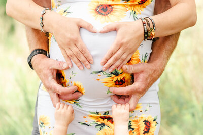 A mother, father, and toddler put their hands around mama's growing belly during a maternity session.