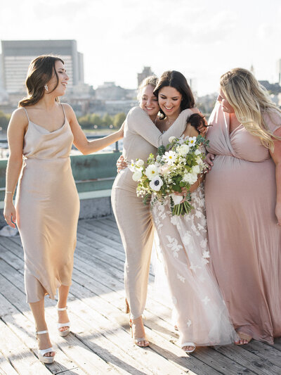 Bridesmaids embrace a bride holding a lush bouquet on the rooftop of the Science Centre at sunset. 