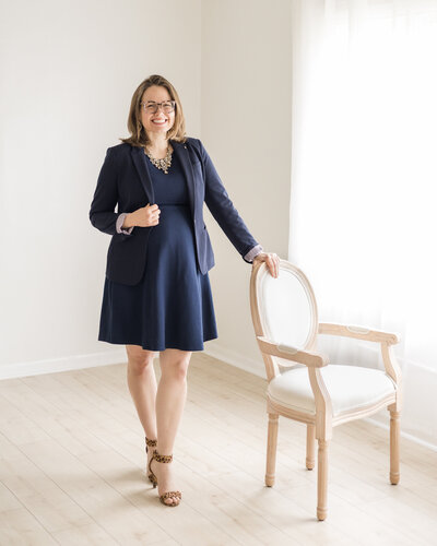 pregnant woman standing in a studio room, next to a chair with her hand on it, facing the camera smiling, wearing a glasses, dress, heels, and a dress jacket,
