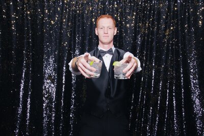 studio lighting photo of a guy holding two drinks posing for a photo booth front of a black sequin backdrop