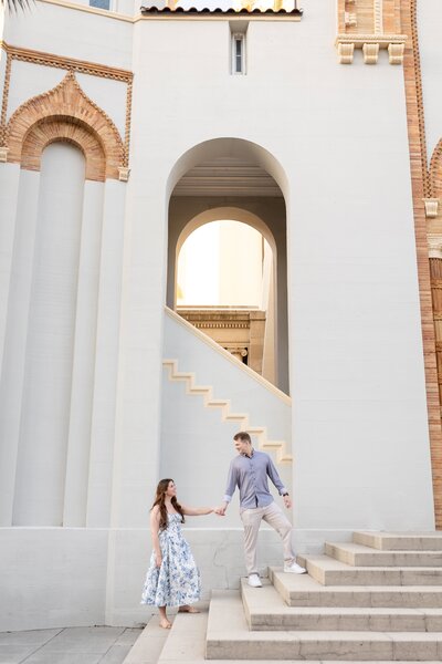  couple shares a romantic moment during their engagement session in St. Augustine, Florida. The man reaches down to help his fiancée up a grand staircase framed by architectural arches and soft sunset light. Her flowy floral dress adds movement to the serene, refined setting. Captured by a Jacksonville engagement photographer who blends documentary storytelling with a touch of elegance and architectural charm.