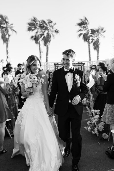 Black-and-white photo of a newlywed couple walking down the aisle, smiling, as guests applaud at an outdoor wedding in Palm Springs, California, with palm trees in the background.