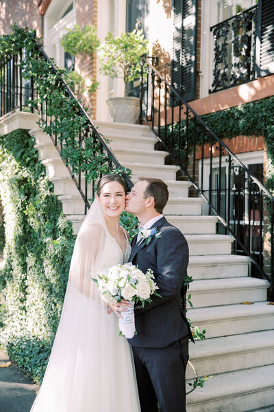 A newlywed couple shares a joyful moment in front of a stately Southern-style home, surrounded by greenery and black iron railings, featured by Grey Cove Designs for timeless wedding storytelling.