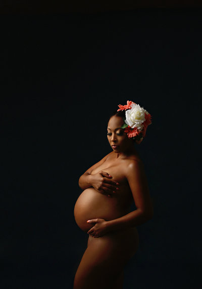 pregnant black woman nude with arm across chest with floral hair piece against black backdrop