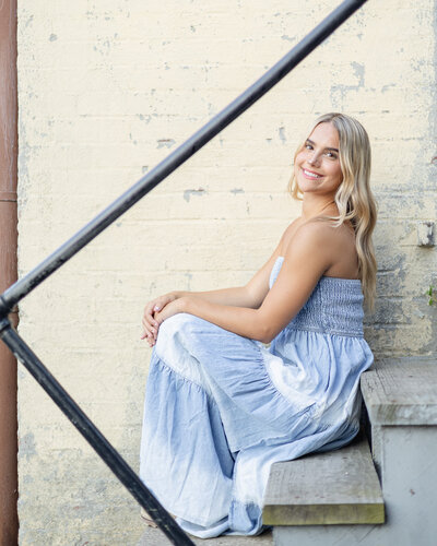high school senior female with long blonde hair, sitting on steps next to a yellow brick wall, wearing a white and blue strapless dress, smiling at the camera