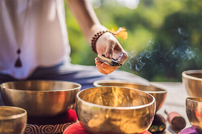 A hand holding burning wood over gold sound healing bowls