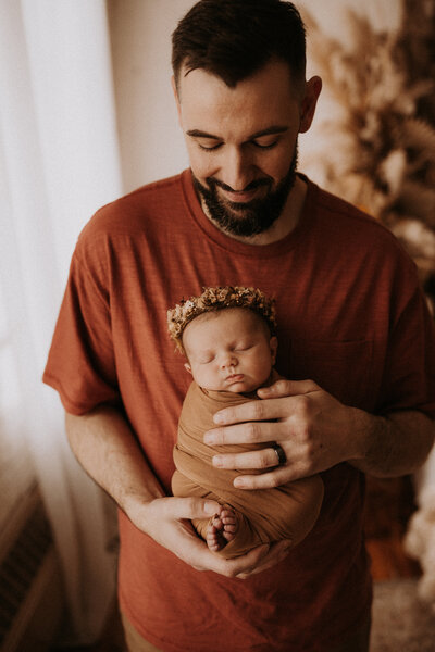 Father holding his newborn baby girl wearing a flower crown and swaddled in a blanket.