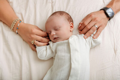 Connecticut newborn photographer captures peaceful sleeping baby in white knit outfit with parents' hands gently touching on cream blanket backdrop