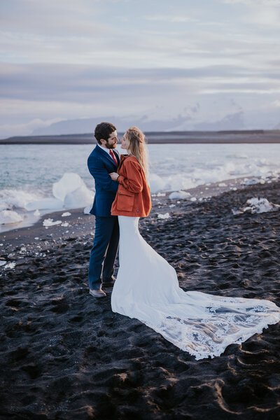 Bride and groom embrace on Iceland’s black sand beach with scattered glacier chunks, a magical scene with an Iceland wedding photographer.