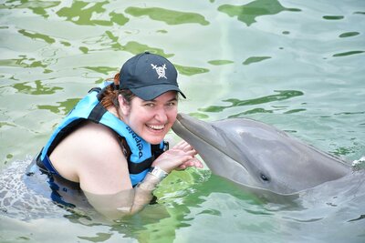 A woman smiles happily up at the camera from the water, wearing a life jacket and a black baseball cap. A dolphin has lifted its face from the water and is gently kissing the woman's cheek.