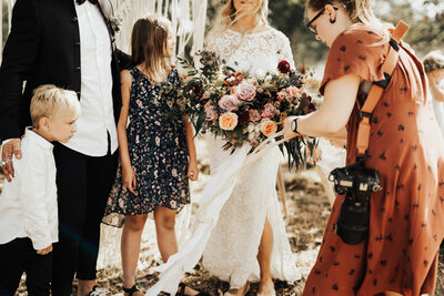 bröllopsfotograf hjälper bruden med bröllopsbuketten inför gruppbild med barn efter vigsel på bergs gård i trosa