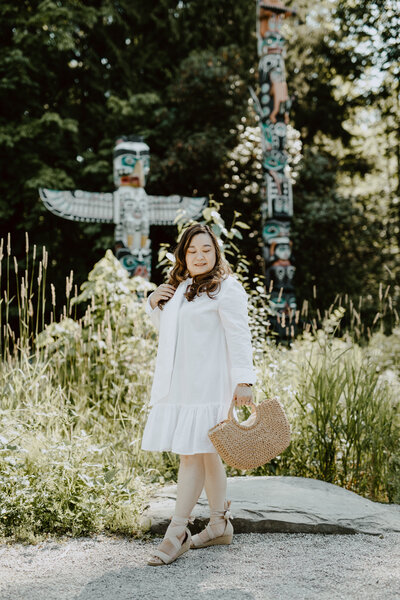 Anne, an Asian woman, is in walking position in front of the Stanley Park Totem Poles in Vancouver, BC, Canada. Her eyes are looking down to the ground as her right hand is brushing her hair. She holds a straw purse in her left hand as she wears a knee length white dress with suede espadrilles.
