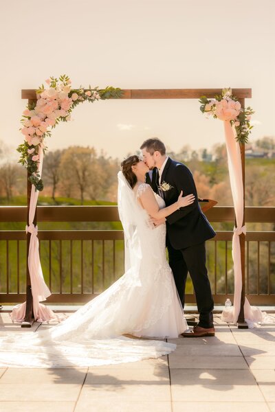 A bride and groom kiss under a floral arch on a patio.