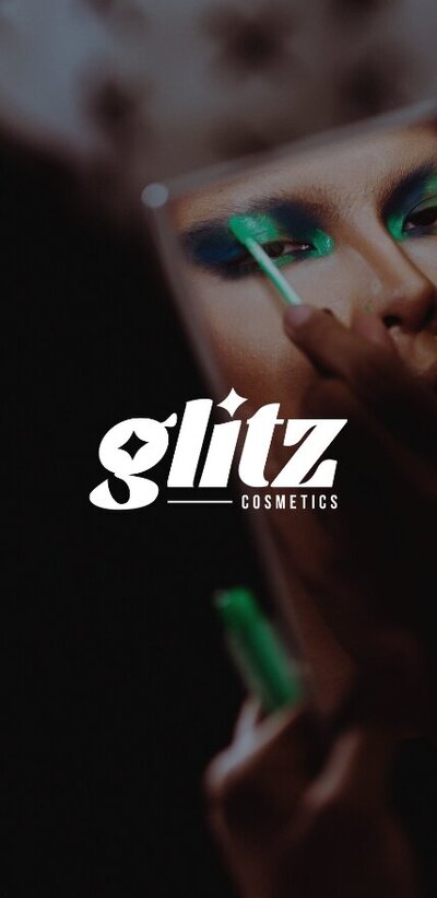 Person putting on green eyeshadow in a handheld mirror with a white logo that reads "Glitz Cosmetics" over the top