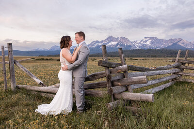 Bride and grrom holding each other face to face with fence and Sawtooth mountains in the back gground.