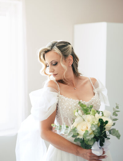 A bride in a white wedding dress holds a delicate bouquet of white and green flowers in a bright, airy setting.