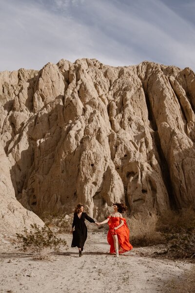 Just married couple celebrating in the desert, one partner popping a champagne bottle while the other laughs and holds a bouquet.