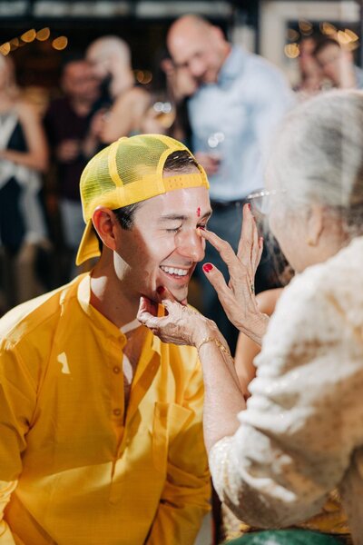 A smiling person in a yellow shirt and baseball cap receives a red mark on the forehead from an older individual's hands during a cultural ceremony.