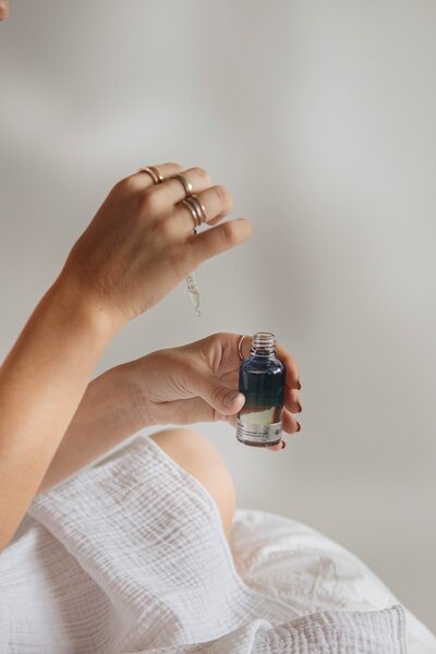 A hand holds a dropper bottle of essential oil, ready for a calming skincare ritual, set against a soft, white blanket for a cozy, relaxing vibe.