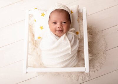 A newborn is peacefully swaddled in soft white fabric, resting on a cozy blanket with star-patterned pillows nearby.