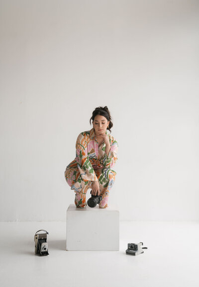 Photographer in a colorful floral jumpsuit squats on a white cube, holding a camera. Two vintage cameras are placed beside her on the floor. Neutral expression.