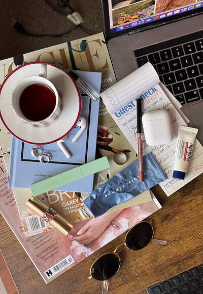 Messy flat lay of a stack of magazines and books, a coffee mug, sunglasses, and lipstick on a table