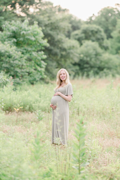 An expecting mother wearing a green dress stands in a field of grasses and smiles.