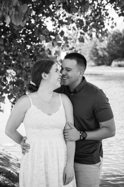 A couple embrace side by side with leaves of a tree behind them. 