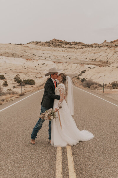 A groom who is holding his bride by the waist and kissing her in the middle of a paved road. There is light beige rock and sagebrush behind them in the distance.