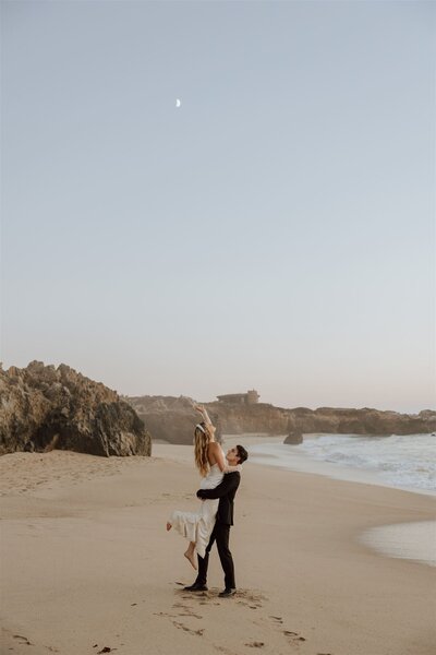groom lifting bride on a beach, bride is reaching for the moon in the sky