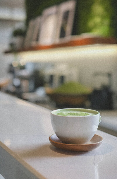 A white cup of matcha latte sits on a brown saucer on a white counter, reminiscent of the serene settings often described in Phillipa Leseberg's books, with blurred shelves in the background.