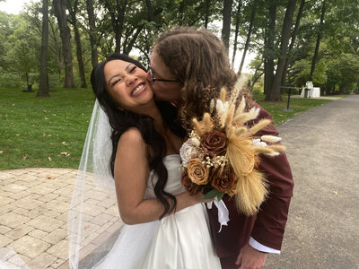 Bride and groom share a kiss in a lush outdoor setting, holding a dried floral bouquet.