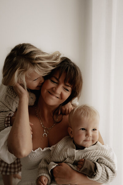 A woman in a beige dress holds a newborn and embraces a young child in a hat, standing in a grassy field with the South Coast ocean in the background, beautifully captured by an NSW family photographer.