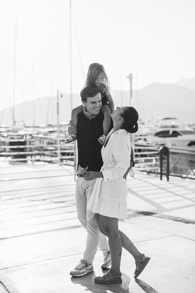Candid black and white photo of a joyful family walking by the marina with their daughter on dad’s shoulders, captured during a sunset lifestyle session