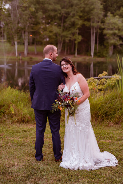 bride hugging groom from the side standing in opposite direction in front of pond with bouquet during summer wedding at avonlea farms coordinated by cait and co events