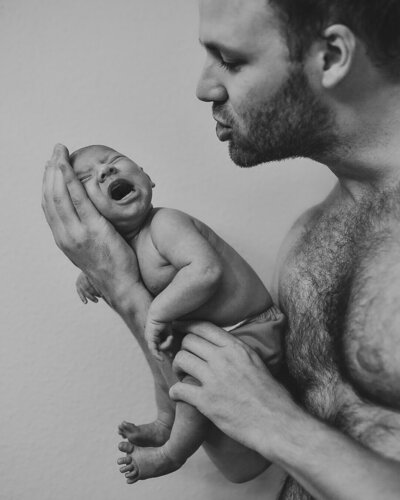 black and white photo of a father holding his crying newborn on his one hand, while shushing  to calm the baby down.