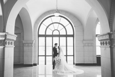 A black and white film photo of a bride and groom against a window at the Majestic in Downtown Los Angeles