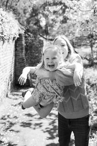 A black and white image capturing a mother's playful flight with her giggling toddler under a canopy of trees, taken by Suzy Slemen Photography.