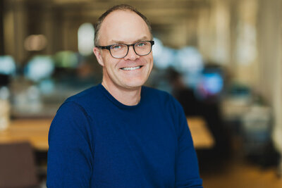 Professional portrait of middle-aged man in blue sweater and glasses, smiling warmly in office background