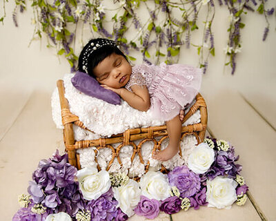 Baby girl wearing a purple lace dress, sleeping on her tummy on a basket with purple flowers around her.