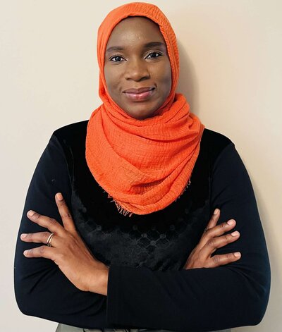 photo of a smiling woman against a beige background. she is wearing an orange headscarf with a black long sleeve shirt and her arms are crossed. 