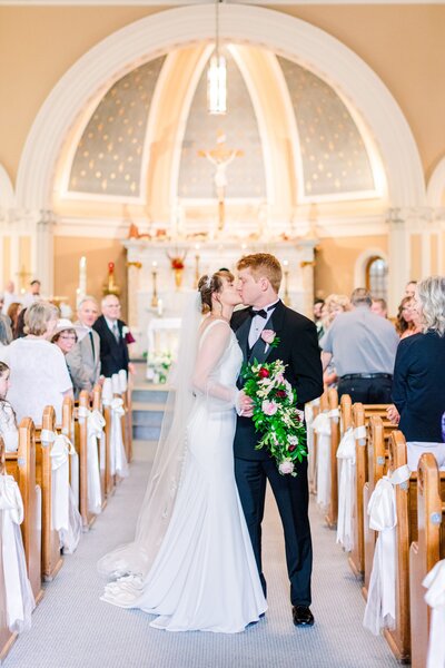 Bride holding a cascading bouquet of red and pink flowers kisses her groom as they walk back up the aisle of their Coeur d'Alene Wedding venue 