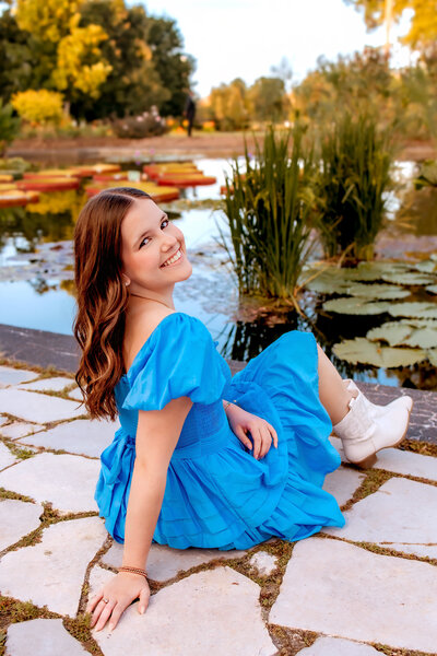 A brown haired girl wearing a white dress and brown cowboy boots poses herself leaning against a stone wall in Tower Grove Park.