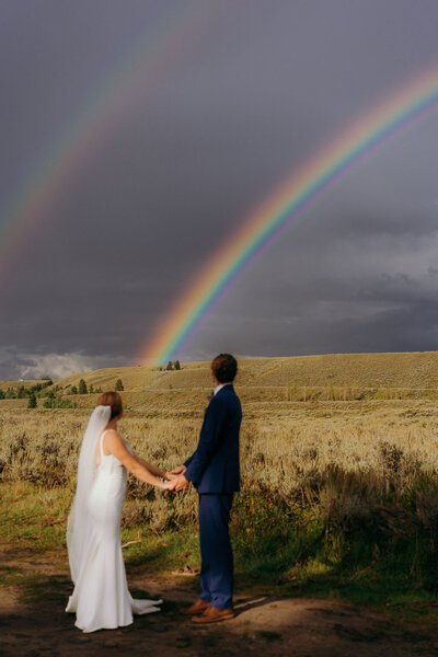 photo of bride and groom holding hands while looking at a double rainbow taken by Grand Teton elopement photographer
