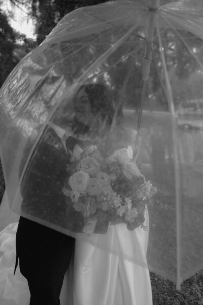 A moody, artistic photo of a bride and groom embracing under a clear plastic umbrella in the rain. The couple is kissing, surrounded by a blurred, rainy outdoor background, creating a cozy and intimate atmosphere.