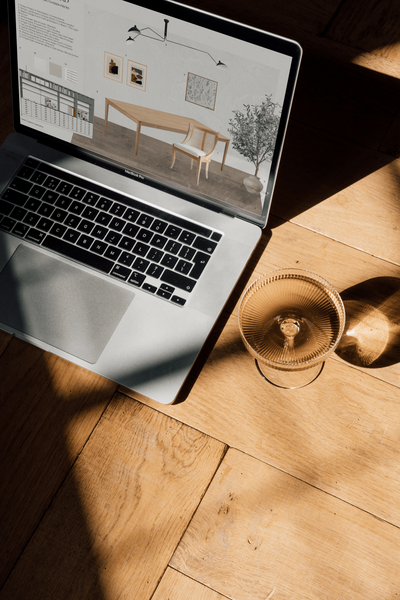 Laptop sitting on herringbone timber flooring, with a champagne coupe filled with rosé beside it. Sunlight streams in, casting beautiful light and shadows. The laptop displays a sample board for a formal meeting room designed for an interior design studio.