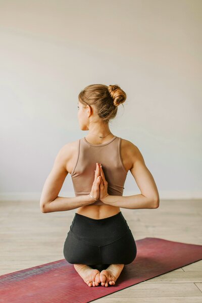 A woman practicing yoga in a minimalist studio. She wears a beige tank top and black high-waisted leggings. Her back faces the camera as she holds a prayer position with her hands behind her back. Her blonde hair is tied in a bun, and she gazes slightly to the left.