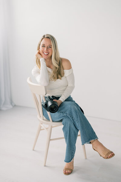 a woman sits in a chair, crosses her legs while holding her camera, and has her chin in her hands as she smiles at the camera. She is wearing blue jeans and a white abercrombie off the shoulder top and sits against a white background